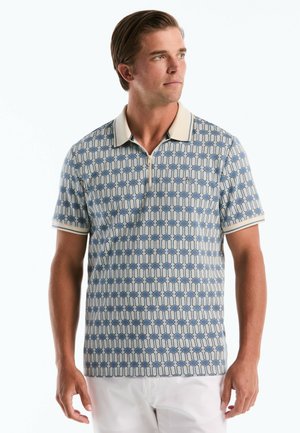 Mann trägt kurzärmeliges blau-beigefarbenes Polohemd mit Muster, weißem Kragen und weißer Hose, schaut zur Seite.