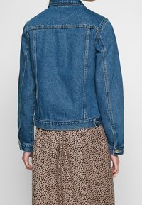 Denim jacket in medium blue with a classic cut, double-stitched seams, and button closures, worn over a patterned skirt.