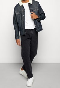 Giacca di jeans con collo in pelliccia, lavaggio scuro, pantaloni grigi e sneakers bianche. La giacca ha chiusura con bottoni e tasche sul petto.