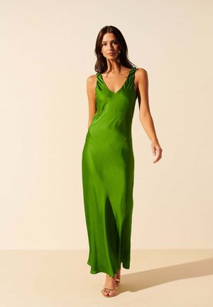 Femme aux longs cheveux bruns portant une robe longue en satin vert sans manches et des talons ouverts beiges, debout devant un fond beige uni.