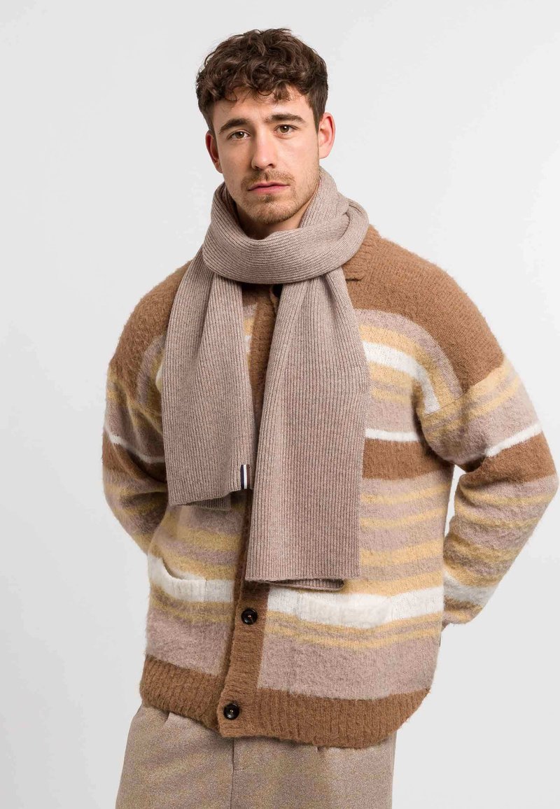 Un jeune homme portant un cardigan rayé marron, beige et blanc et une écharpe tricot beige, se tenant contre un fond clair uni.