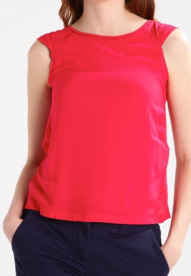 Femme portant un débardeur rouge vif et un pantalon bleu foncé, debout les mains dans les poches devant un fond uni.