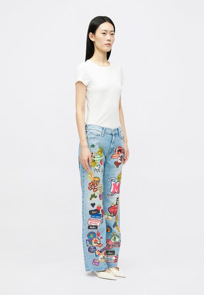 Femeie stand în top alb cu mâneci scurte și blugi deschiși la culoare, acoperiți cu petice colorate și text, purtând pantofi albaștri cu vârf ascuțit.