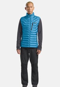 Gilet puffer leggero blu con baffle orizzontali e tasca sul petto con zip. Indossato sopra una maglietta a maniche lunghe scura e abbinato a pantaloni neri.