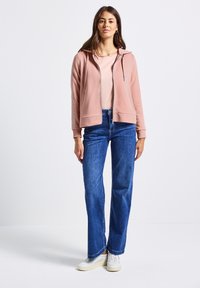 Felpa rosa con cappuccio e zip, dal texture morbido, abbinata a una t-shirt chiara e a jeans larghi blu. Sneakers bianche completano l'outfit.