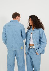 Jachete din denim de culoare albastră deschisă, cu cusături vizibile, croială lejeră și închidere cu nasturi. O jachetă are o patch cu logo, în timp ce cealaltă are broderie.