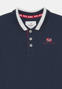 Námořnická polo košile s bílým a červeným límečkem, třemi knoflíky a červeným vyšitým logem na levém prsu. Hladký bavlněný materiál.