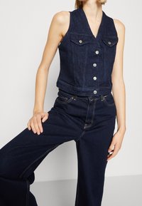 Kamizelka jeansowa w ciemnym niebieskim kolorze, bez rękawów, z głębokim dekoltem w serek, dwoma kieszeniami na piersi i metalowymi guzikami z przodu. Dopasowana do ciemnych jeansów w tym samym kolorze.