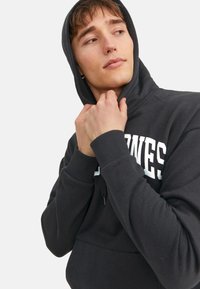 Dunkelgrauer Kapuzenpullover mit einer Fronttasche, gerippten Bündchen und Kapuze mit Kordelzug. Der Text erscheint in fetten weißen Buchstaben auf der Brust.