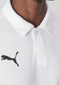 Polo blanc en tissu texturé, col pointu, fermeture à boutons sur le devant, avec un logo noir représentant un chat sautant sur la poitrine.