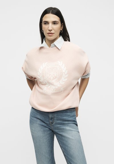 Femme aux longs cheveux foncés portant des boucles d’oreilles créoles, un sweat-shirt rose pâle avec un logo blanc, une chemise blanche rayée à col en dessous, et un jean bleu.