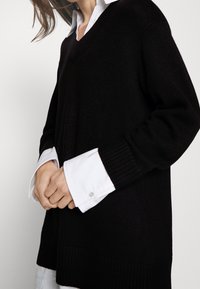 Maglione nero a collo a V in maglia con polsini a coste, indossato sopra una camicia bianca con colletto. La texture sembra morbida, con un design a vestibilità ampia e rilassata.