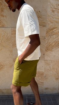 T-shirt bianco in cotone abbinato a pantaloni corti verde oliva, con tasche e un bordo nero all'orlo. Lo sfondo mostra un muro di pietra testurizzato.