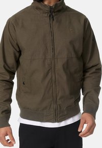 Veste légère vert olive avec fermeture éclair à l'avant, poignets côtelés et deux poches latérales. Texture lisse avec un logo discret sur la poitrine.