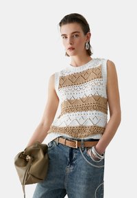 Haut sans manches en maille avec motifs rayés blancs et beige, design ajouré et encolure côtelée, associé à un jean bleu et une ceinture marron.