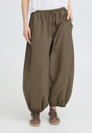 Pantaloni larghi verde oliva con vita e polsini elasticizzati, indossati con sneaker marroni e top bianco, mani parzialmente nelle tasche.