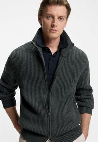 Mörkgrå stickad cardigan med hög krage, ribbad textur, dragkedja fram och rullade ärmslut, lagd över en marinblå polo skjorta.