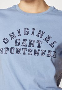 T-shirt in cotone azzurro chiaro con una stampa di testo curva in blu navy che legge "ORIGINAL GANT SPORTSWEAR" sulla parte anteriore, in diverse dimensioni di carattere.