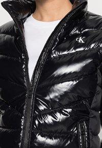 Czarna kurtka puchowa o błyszczącej, pikowanej powierzchni, z wysokim kołnierzem i pełnym zamkiem błyskawicznym. Posiada małe logo na klatce piersiowej i boczne kieszenie.