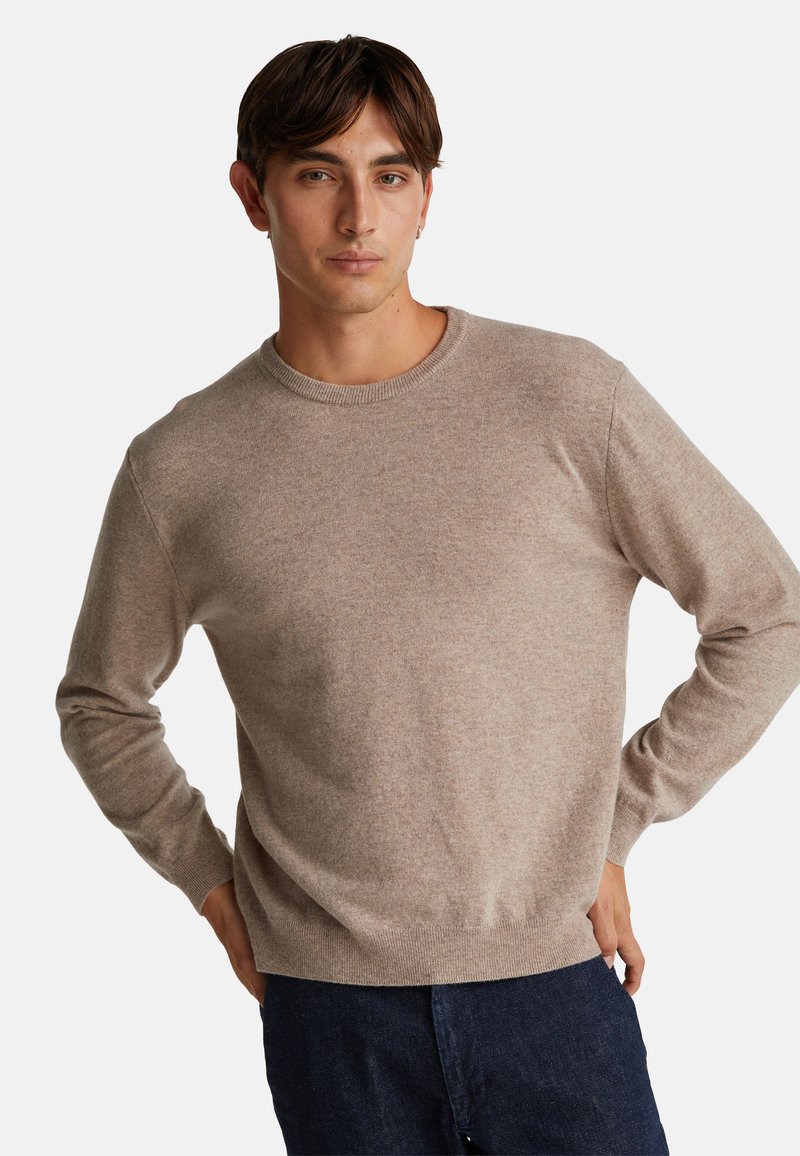 Maglione beige a maglia con scollatura a girocollo, polsini e bordo a costine. Il modello posa davanti a uno sfondo semplice, indossando pantaloni in denim scuri.