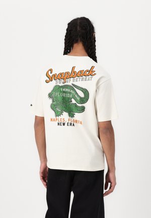 Crème graphic t-shirt met een groen alligator ontwerp en de tekst "Snapback Swamp Retreat" en "Florida" in oranje en zwart.