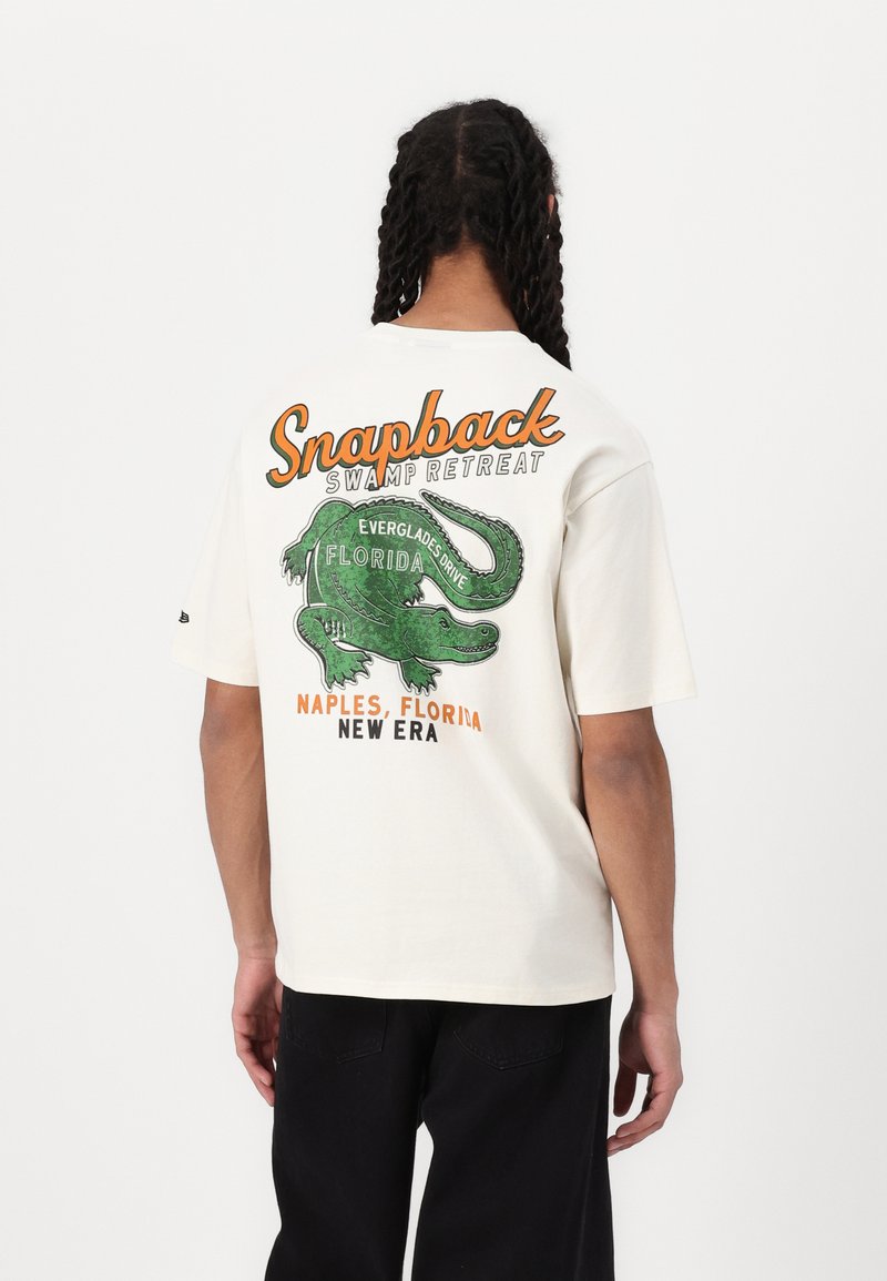 Cremefarvet grafisk t-shirt med et grønt alligator design og teksten "Snapback Swamp Retreat" samt "Florida" i orange og sort.
