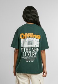 Miško žalios spalvos T-shirt su laisvu stiliumi. Nugaros pusėje yra didelis baltas ir oranžinis grafikas su tekstu: "Offline YRA NAUJAS PRABANGOS STANDARDAS" virš retro televizorių atvaizdų.
