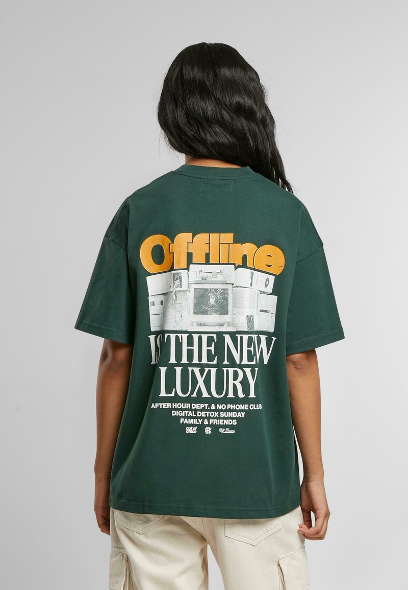 Miško žalios spalvos T-shirt su laisvu stiliumi. Nugaros pusėje yra didelis baltas ir oranžinis grafikas su tekstu: "Offline YRA NAUJAS PRABANGOS STANDARDAS" virš retro televizorių atvaizdų.