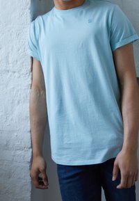 Ljusblå t-shirt med korta ärmar, tillverkad av mjuk bomull, med uppvikta ärmslut och en minimalistisk logotyp på bröstet, i kombination med mörka jeans.