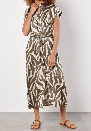 Vrouw draagt een midi-jurk met korte mouwen, knopen en een ceintuur, met een olijfkleurige en witte abstracte zebra-print en bruine platte sandalen.