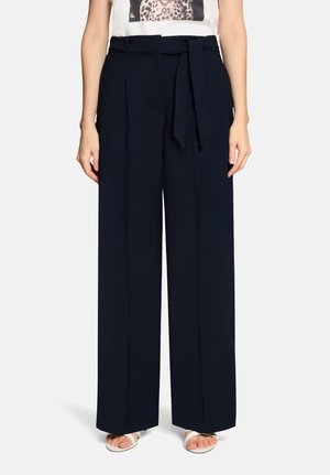 Kobieta w granatowych, szerokich spodni z wysokim stanem, przewiązanych paskiem, oraz białych sandałach na odkryte palce, na jednolitym tle.