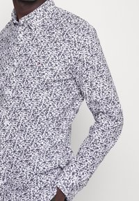 Valkoinen pitkähihaisessa paidassa on tummansininen ja harmaa abstrakti kuvio, jossa on nappikaulus ja hienovarainen logotunnus rinnassa.