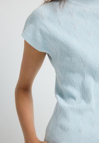 Kvinna som bär en ljusblå kortärmad stickad topp med texturerat bladmönster, synlig från midjan till överarmen mot en enfärgad bakgrund.
