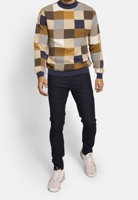 Pull en tricot avec un motif géométrique marron, crème et bleu marine ; encolure arrondie et poignets côtelés, associé à un jean en denim sombre et des baskets.