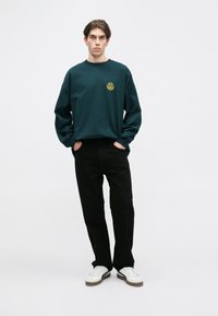 Dark green long-sleeve t-shirt with a round neckline and a yellow logo. Paired with black pants and white sneakers. Minimalist design.
