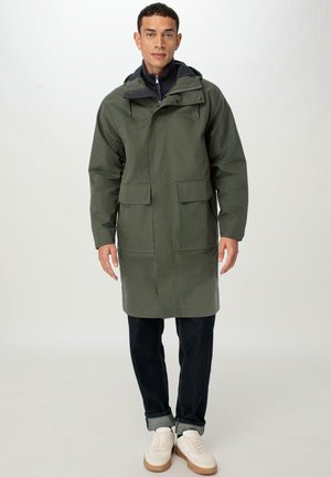 Olive green raincoat with a hood, featuring a front zipper, two large front pockets, and a straight cut, paired with dark jeans and white sneakers.