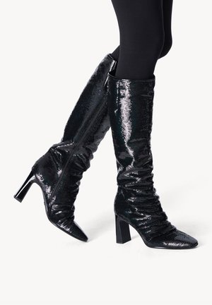 Bottes noires montantes en matériau pailleté brillant avec un design affaissé, bouts pointus et talons épais. Comprend une fermeture éclair sur le côté.