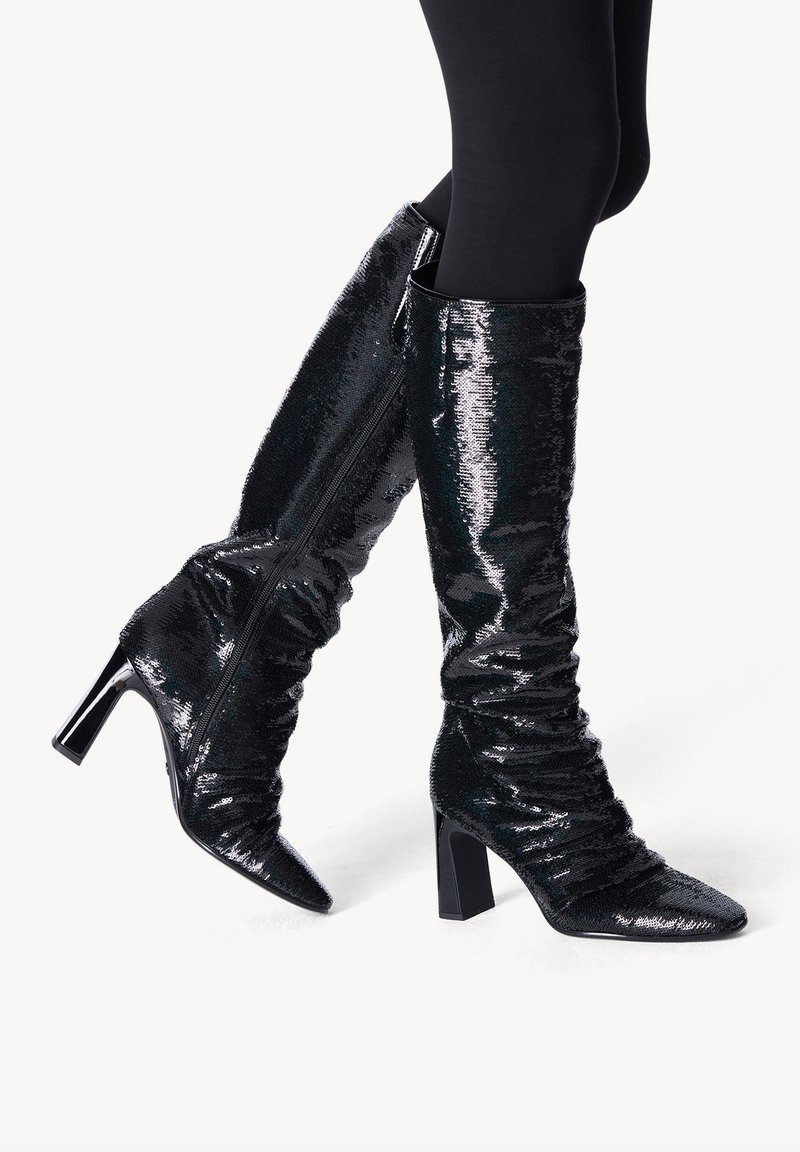 Botas negras hasta la rodilla hechas de un material brillante de lentejuelas con un diseño arrugado, punteras afiladas y tacones gruesos. Presentan una cremallera lateral.