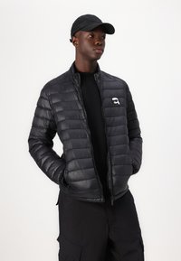 Schwarze, aufgeplusterte Jacke mit horizontaler Steppstruktur, Reißverschluss und Logo-Akzent. Trägt dazu ein schwarzes Hemd und schwarze Hose.