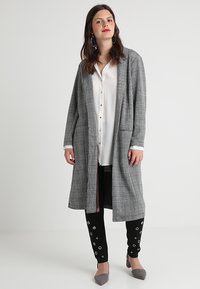 Szürke kockás hosszú kabát laza szabással, két elülső zsebbel és kontrasztos piros béléssel. Fekete nadrággal és szürke magassarkúval párosítva.