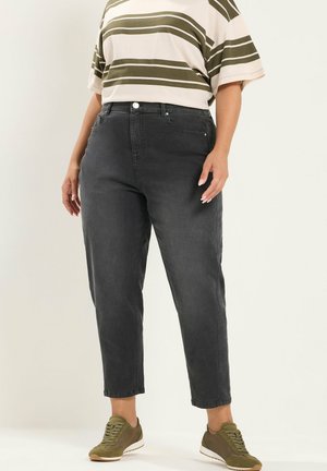 Person trägt schwarze High-Waist Slim Jeans, grün-weiß gestreiftes kurzärmeliges Shirt und olivgrüne Sneakers und steht vor weißem Hintergrund.