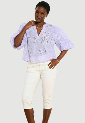 Žena v ľaliovej blúzke s nafúknutými rukávmi s kvetinovými výrezmi a bielych krátkych nohaviciach, pózujúca s jednou rukou na bokoch a druhou pri krku.