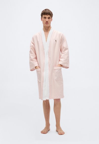 Νεαρός άνδρας που στέκεται ξυπόλυτος φορώντας μία ρόμπα με ριγέ μοτίβο σε ανοιχτό ροζ και λευκό, με τσέπες και μικρό κεντημένο λογότυπο στο στήθος.