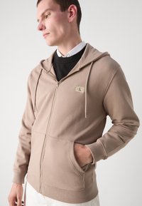 Felpa con zip beige realizzata in tessuto morbido, dotata di tasca a marsupio, polsini a costine e una piccola etichetta del logo sul petto.