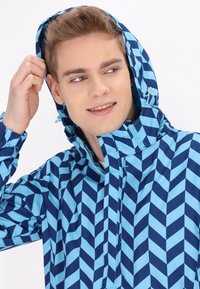 Svetlo modra in modra zigzag vzorec jakna s kapuco, spredaj na zadrgo in elastičnimi manšetami, ki prikazuje lahkotno, vodoodporno prevleko.