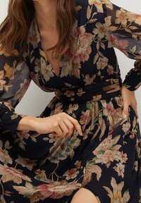 Woman wearing a dark floral long-sleeve dress with a V-neck and a wide black belt, holding the skirt with one hand.