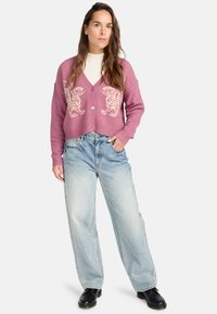 Cardigan en tricot rose avec motif de tigre, boutonné à l'avant et au niveau des poignets côtelés, associé à un jean large bleu clair et des bottines noires.