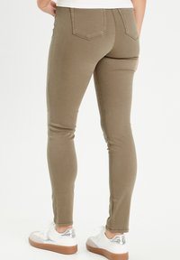 Jeans extensibles vert olive avec une coupe ajustée, deux poches arrière et des détails de couture visibles. Associés à des baskets blanches avec des accents en argent.