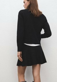 Outfit composto da un maglione nero a maglia e una gonna svasata con maniche lunghe. La gonna ha un alto punto vita e una superficie liscia, sovrapposta a una maglietta bianca.