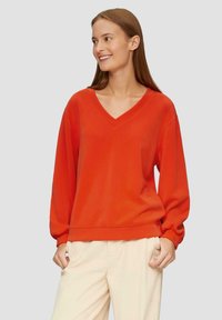 Czerwony sweatshirt z dekoltem w serek, z długimi, marszczonymi rękawami i luźnym krojem. Gładka faktura, ściągacz u dołu i przy mankietach. Noszony z jasnymi spodniami.